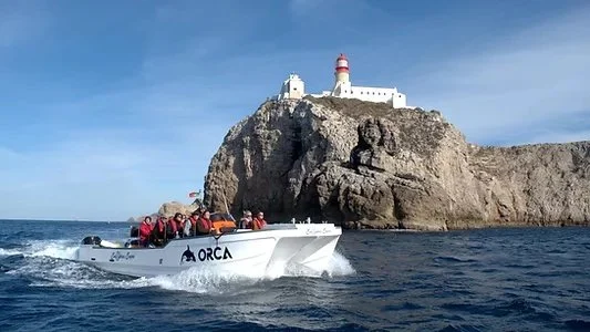 Boat named Orca cruising below a cliff-top lighthouse