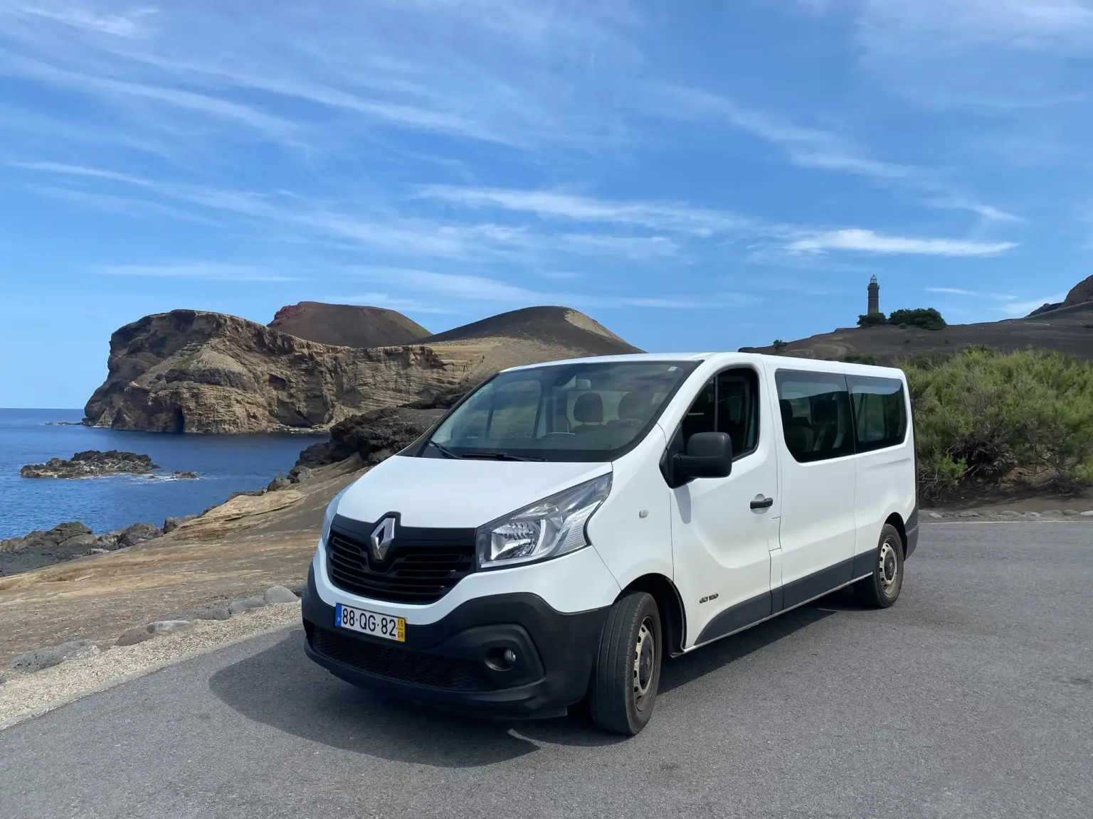 White minibus parked at a coastal viewpoint with a lighthouse and headland behind