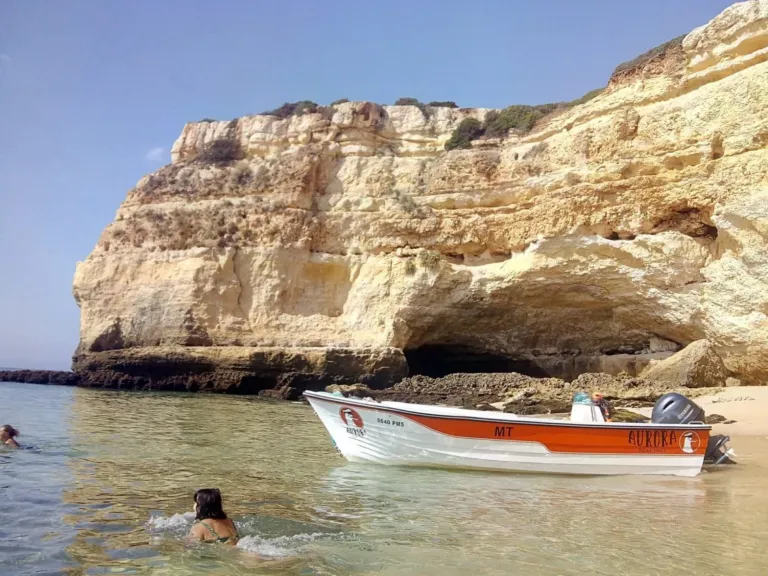 Small orange-and-white boat anchored below an eroded sandstone cliff, with a swimmer nearby