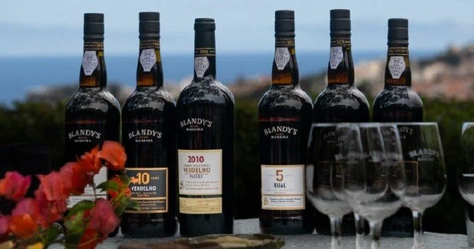 Four bottles of aged Madeira wine and glasses on a table with flowers and the sea behind
