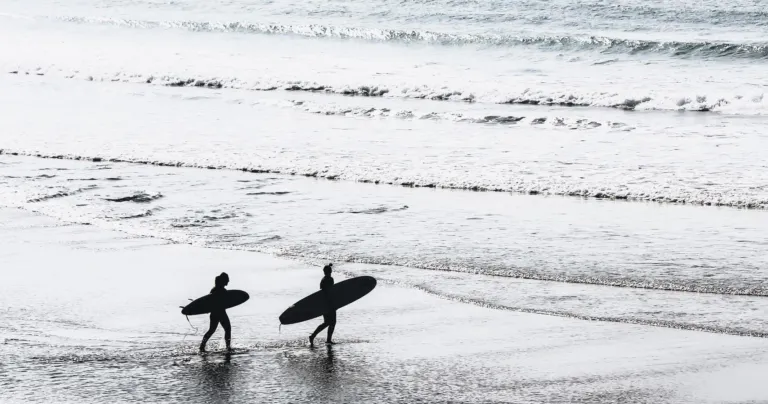 Two surfers carrying boards along a wet beach in monochrome light