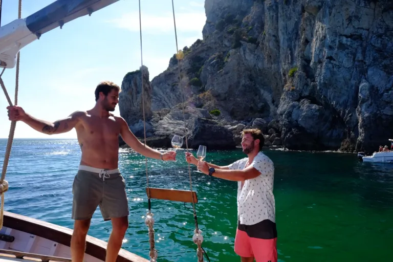 Two men toasting with glasses on a sailing boat in a rocky bay