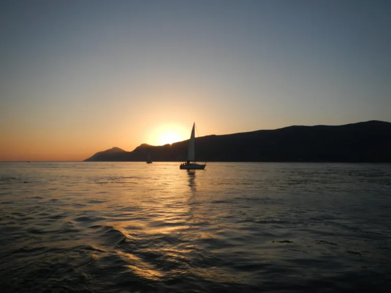 Sailboat silhouetted against the setting sun beside a dark headland